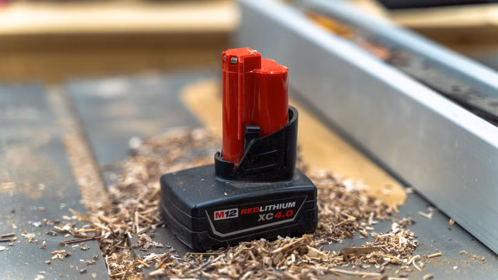 A Milwaukee M12 battery sitting on a table saw with sawdust around it.