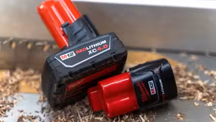 Two Milwaukee M12 batteries sitting on a table saw with sawdust and a blade in the background.