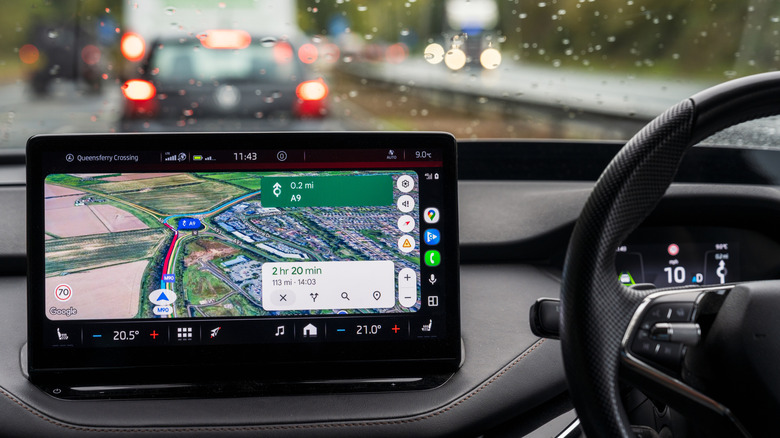 An in-car infotainment display shows a Google Maps navigation route while the vehicle is being driven in rainy traffic.