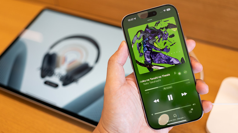 Hand holding an iPhone with Apple Music open to a Now Playing screen, with an iPad blurred in the background.