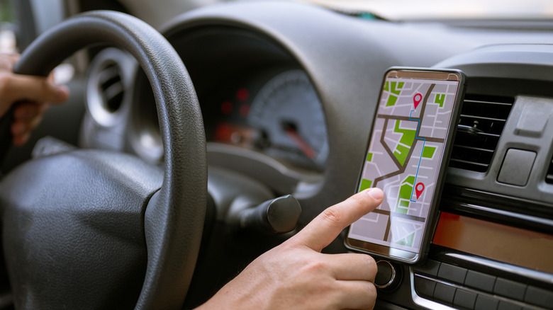 A smartphone mounted beside a car's dashboard displays turn-by-turn navigation while a driver reaches toward the screen.