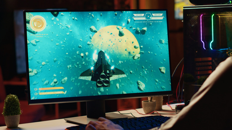 Person playing a space game on a desktop monitor beside an RGB-lit gaming PC.