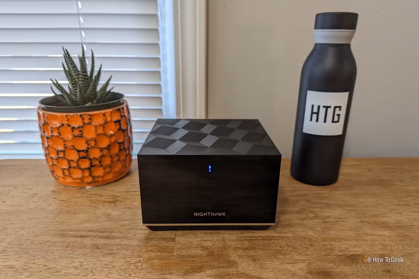 A Netgear Nighthawk MK93S mesh node on a table between a plant in an orange pot and a black water bottle.