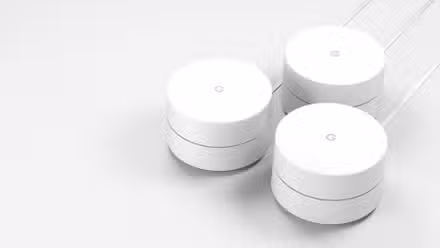 Three white cylindrical mesh Wi-Fi router units arranged together on a light surface, each with a subtle “G” logo on top.